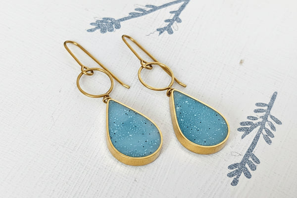 A pair of silver drop earrings with teardrop-shaped light blue speckled resin pendants, displayed on a white textured surface with a blue botanical print.