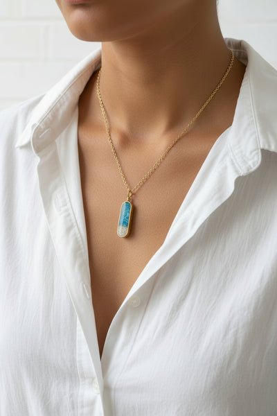 A gold chain necklace with a horizontally oriented capsule pendant, featuring a blue and white marbled pattern on one half and white glitter on the other, displayed on a white geometric stand.