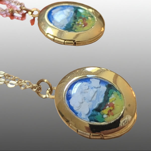 Gold locket necklace with a colorful landscape painting insert, shown on a light wood surface with another locket in the background.